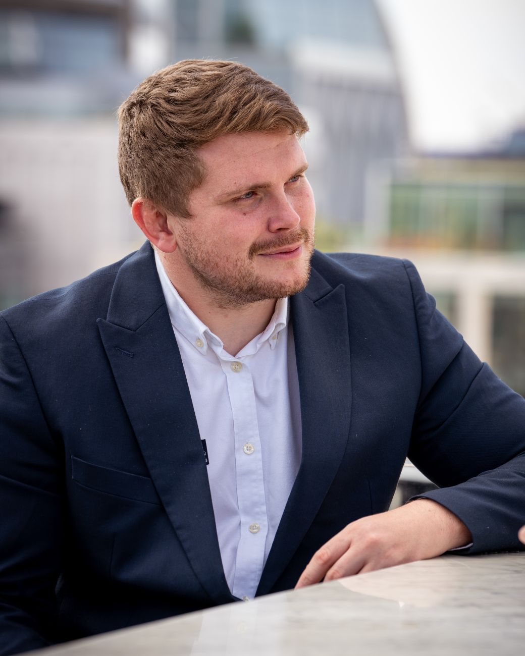 A man with short hair and a beard, wearing a navy blue blazer and white shirt, is sitting outdoors against a blurred urban backdrop. The confident demeanor of this property sourcing expert suggests he's well-versed in his field.