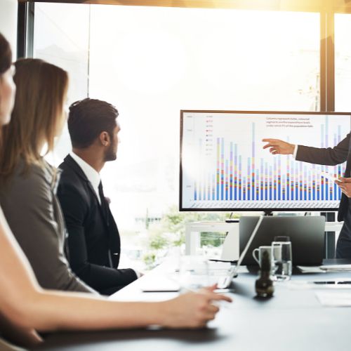 A businessperson from a renowned Business Growth Consultancy presents a bar graph on a large screen to three colleagues in a modern conference room.